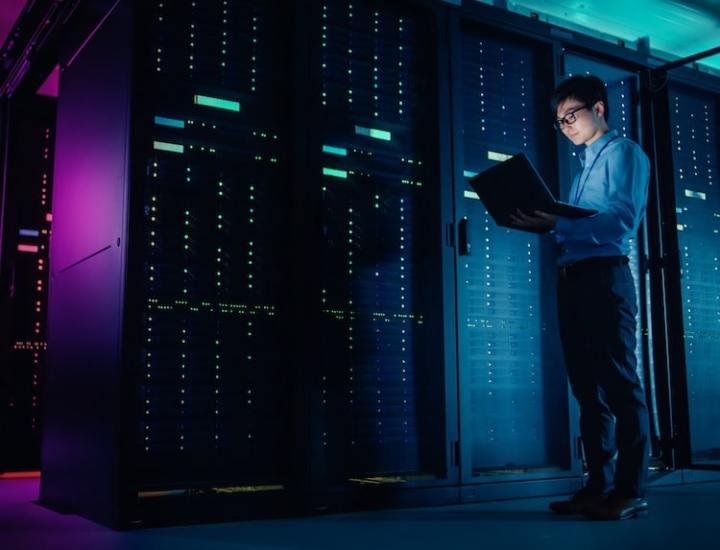 IT technician standing in a data center holding a laptop, surrounded by server racks