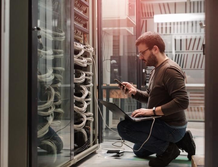 IT technician kneeling on the floor in a server room, working from a laptop and holding a smartphone