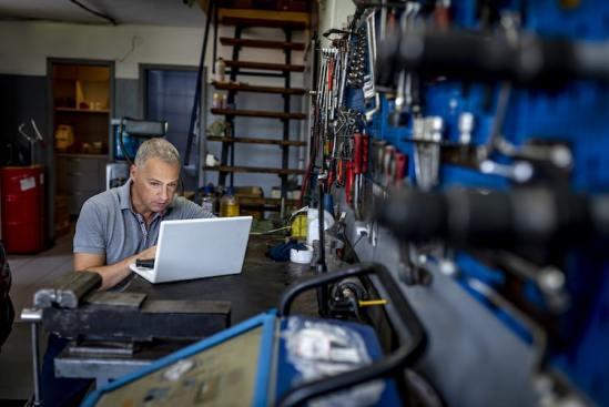 Man working on a laptop in a workshop, surrounded by tools and equipment