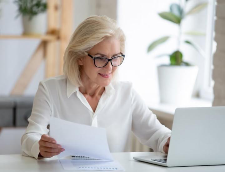 Smiling senior woman with glasses working on a laptop while holding documents in an office