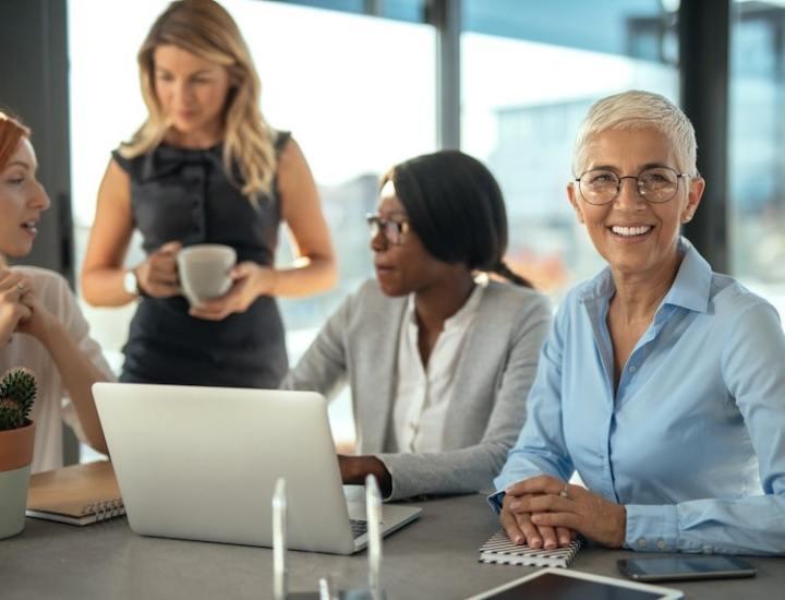 Professional women having a meeting in an office, with one woman smiling at the camera and others engaged in conversation